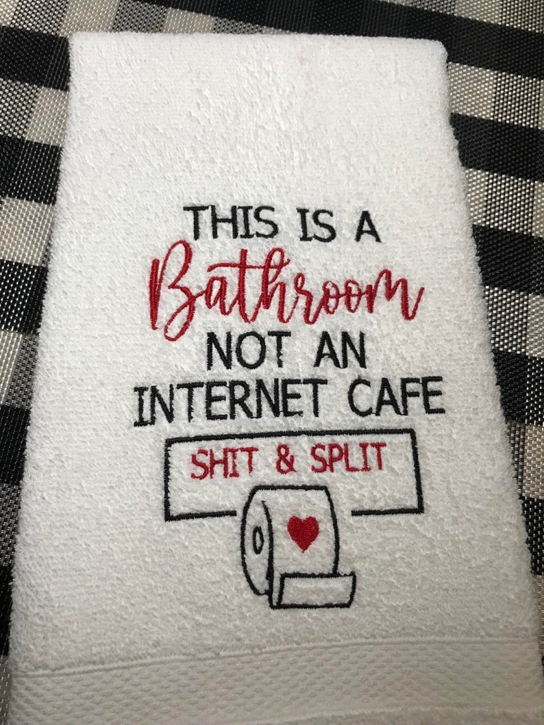 K&ouml;nnte beinhalten: Wei&szlig;es Badezimmer-Handtuch mit gesticktem Text: "THIS IS A Bathroom NOT AN INTERNET CAFE SHIT & SPLIT" und einer Toilettenpapierrolle mit rotem Herz. Das Handtuch liegt auf einer schwarz-wei&szlig; karierten Oberfl&auml;che.