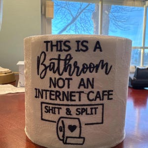 K&ouml;nnte beinhalten: Wei&szlig;e Toilettenpapierrolle mit schwarzem Text: "THIS IS A BATHROOM NOT AN INTERNET CAFE SHIT & SPLIT". Die Rolle hat ein Herzdesign.
