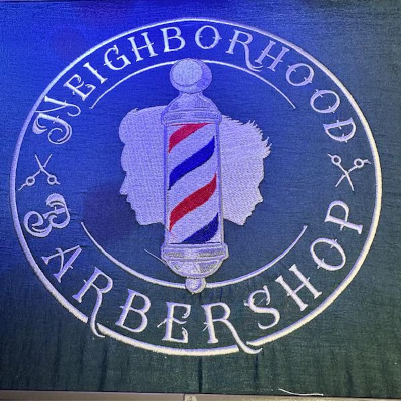 May include: Embroidered sign for a barbershop, featuring a barber pole with red, white, and blue stripes. The sign is circular, with the words "Neighborhood Barbershop" in a white, stylized font. Silhouettes of two faces and scissors are also included.