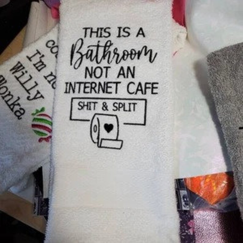 K&ouml;nnte beinhalten: Wei&szlig;es Badezimmer-Handtuch mit dem Text "THIS IS A Bathroom NOT AN INTERNET CAFE SHIT & SPLIT" in Schwarz. Das Handtuch zeigt auch eine Toilettenpapierrollen-Grafik. Andere Handt&uuml;cher sind teilweise sichtbar.