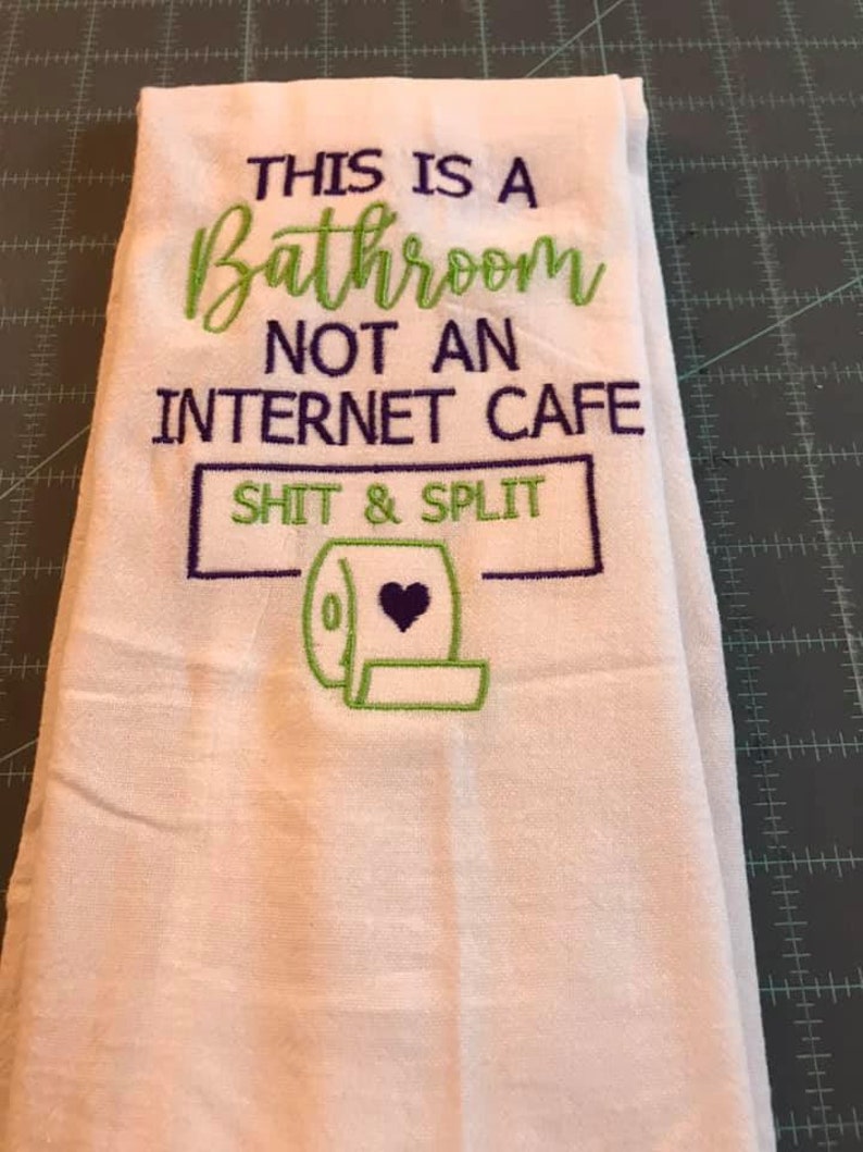 K&ouml;nnte beinhalten: Wei&szlig;es Badehandtuch mit gesticktem Text in Gr&uuml;n und Lila. Der Text lautet "THIS IS A BATHROOM NOT AN INTERNET CAFE SHIT & SPLIT" mit einer Toilettenpapierrollen-Grafik.