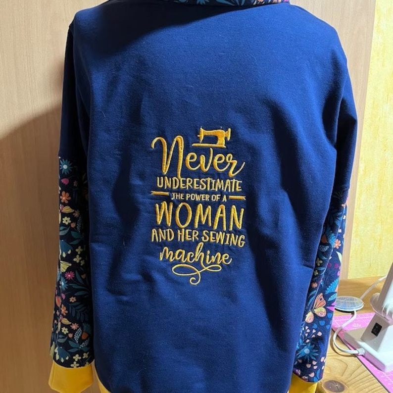 Op de afbeelding: Een marineblauw sweatshirt met mouwen met bloemenprint. De achterkant van het sweatshirt heeft gouden borduurwerk met de tekst "Never underestimate the power of a woman and her sewing machine".