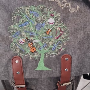May include: A grey canvas backpack with brown leather straps and a large embroidered design of a tree with musical notes and instruments. The tree is green and the instruments are blue, red, and yellow.