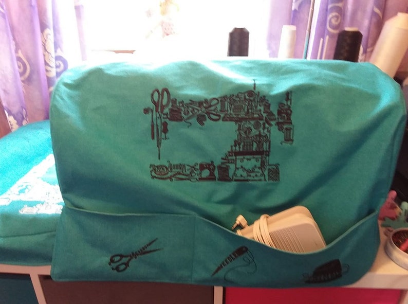 Puede incluir: Funda de m&aacute;quina de coser turquesa con una ilustraci&oacute;n negra de una m&aacute;quina de coser y herramientas de costura. La funda tiene un bolsillo con gr&aacute;ficos de tijeras, aguja e hilo. Se ve un cable de alimentaci&oacute;n de la m&aacute;quina de coser.