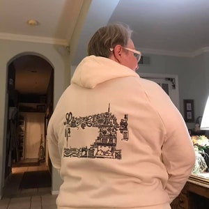 Puede incluir: Sudadera con capucha color crema con un gr&aacute;fico negro de una m&aacute;quina de coser y herramientas de costura. El dise&ntilde;o est&aacute; en la parte posterior de la sudadera. La sudadera tiene capucha y un ajuste relajado.