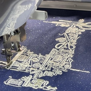 Puede incluir: Una m&aacute;quina de bordar en acci&oacute;n, cosiendo un dise&ntilde;o detallado sobre tela azul marino. El dise&ntilde;o presenta varias herramientas y equipos de costura en color beige claro, creando un efecto texturizado. La tela tiene un aspecto sutilmente brillante.