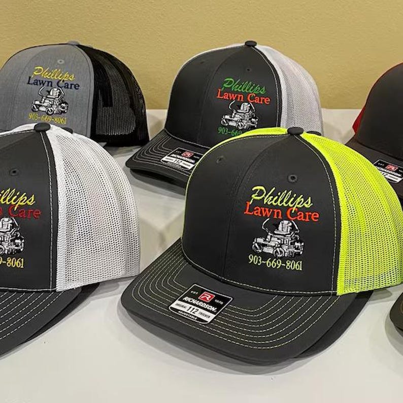 May include: A collection of baseball caps with the "Phillips Lawn Care" logo embroidered on the front. The caps feature a variety of colors, including black, gray, and white, with mesh backs and curved brims. The phone number 903-669-8061 is also displayed.