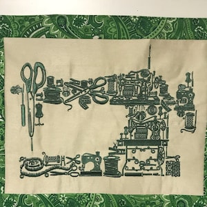 Puede incluir: Un tapiz de tela beige con un borde verde con estampado de cachemira. El centro presenta un dise&ntilde;o bordado de herramientas de costura y m&aacute;quinas de coser antiguas en tonos verdes. Incluye tijeras, carretes y un maniqu&iacute; de sastre.