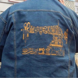 Puede incluir: Chaqueta vaquera azul con un dise&ntilde;o dorado de una m&aacute;quina de coser y herramientas de costura en la espalda. El dise&ntilde;o incluye tijeras, carretes de hilo y una m&aacute;quina de coser. La chaqueta tiene un estilo cl&aacute;sico vaquero.