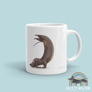 May include: White ceramic mug featuring a whimsical illustration of a dachshund in a yoga pose. The dog is brown with lighter brown spots. The mug has a curved handle and the text "MEET WILMA" is printed on the bottom.