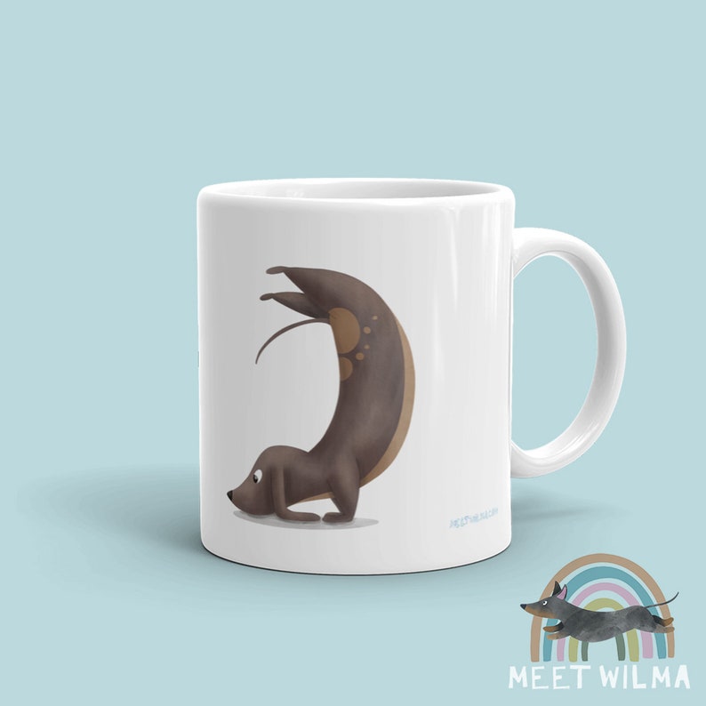 May include: White ceramic mug featuring a whimsical illustration of a dachshund in a yoga pose. The dog is brown with lighter brown spots. The mug has a curved handle and the text "MEET WILMA" is printed on the bottom.