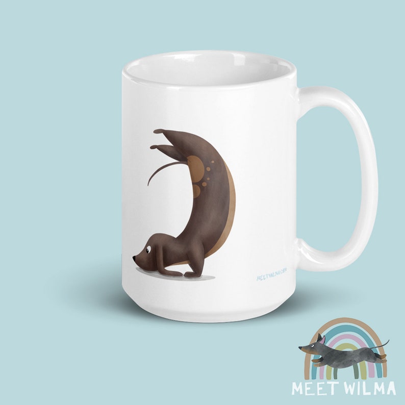 May include: White ceramic mug featuring a whimsical illustration of a dachshund dog in a yoga pose. The dog is brown with lighter brown spots. The mug has a curved handle and the text "MEET WILMA" is printed on the bottom.