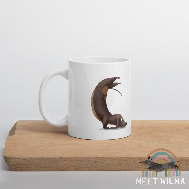 May include: White ceramic mug featuring a whimsical illustration of a brown dachshund dog in a yoga pose. The dog is arched, with its tail and legs extended. The mug has a curved handle and is set on a wooden surface.