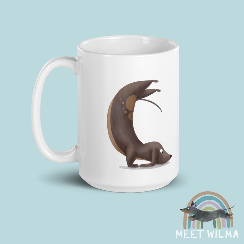 May include: White ceramic mug featuring a whimsical illustration of a dachshund in a yoga pose. The dog is brown with lighter brown spots. The mug has a curved handle and the text "MEET WILMA" is at the bottom.