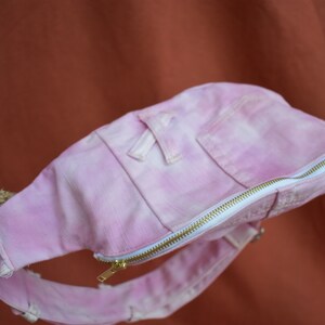 May include: A pink tie-dye denim fanny pack with a white zipper and a small pocket on the front.
