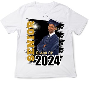 May include: White t-shirt featuring a graduation theme. The shirt has the word "SENIOR" in gold, the text "CLASS OF 2024" in black, and a photo of a graduate in a cap and gown. The background has black brush strokes.