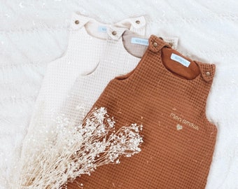 Puede incluir: Dos sacos de dormir para bebé de tejido tipo gofre, uno en beige y otro en marrón. El saco de dormir marrón tiene el texto "Mon amour" bordado en la parte delantera.