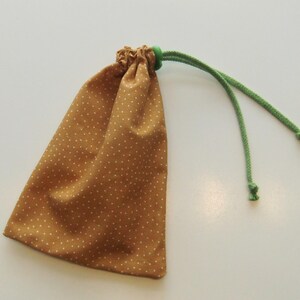 Puede incluir: Una pequeña bolsa con cordón hecha de tela marrón con lunares blancos. La bolsa tiene una parte superior fruncida con un anillo verde y cordones verdes. La bolsa está diseñada para guardar objetos pequeños.
