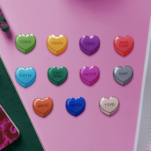 Peut inclure: Neuf cœurs en forme de bonbons de conversation avec des couleurs et du texte différents sur chacun. Les cœurs sont disposés en demi-cercle sur une surface rose. Le texte sur les cœurs est le suivant : TOMG, YBWMM, TSOU, WAN EGBT, OOTW, LWY MMD, YNTCD, TIMT, NBNC, YOYOK, TTPD.