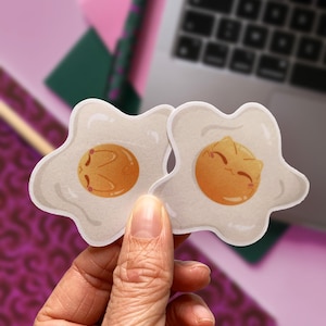 May include: Two white stickers shaped like fried eggs with cute cartoon faces. The stickers are held in a hand.