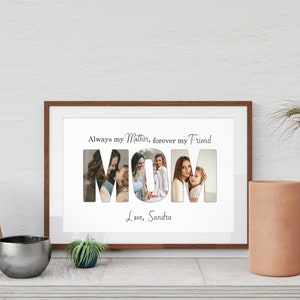 May include: A framed print with the word "MOM" spelled out with photos inside each letter. The text above reads "Always my Mother, forever my Friend" and below "Love, Sandra". The frame is brown and sits on a white surface.