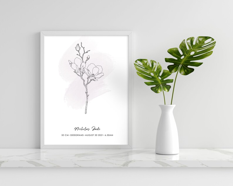 Puede incluir: Una impresi&oacute;n enmarcada con una ilustraci&oacute;n floral en blanco y negro de una rama de magnolia y un fondo rosa acuarela. La impresi&oacute;n incluye el nombre "Nicholas Jade", medidas y fecha. Un jarr&oacute;n blanco exhibe hojas tropicales verdes.