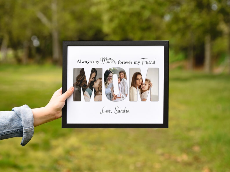 May include: A black framed photo collage with the word "MOM" cut out to reveal four family photos. The text above reads "Always my Mother, forever my Friend" and below "Love, Sandra". The frame is held against a blurred green background.