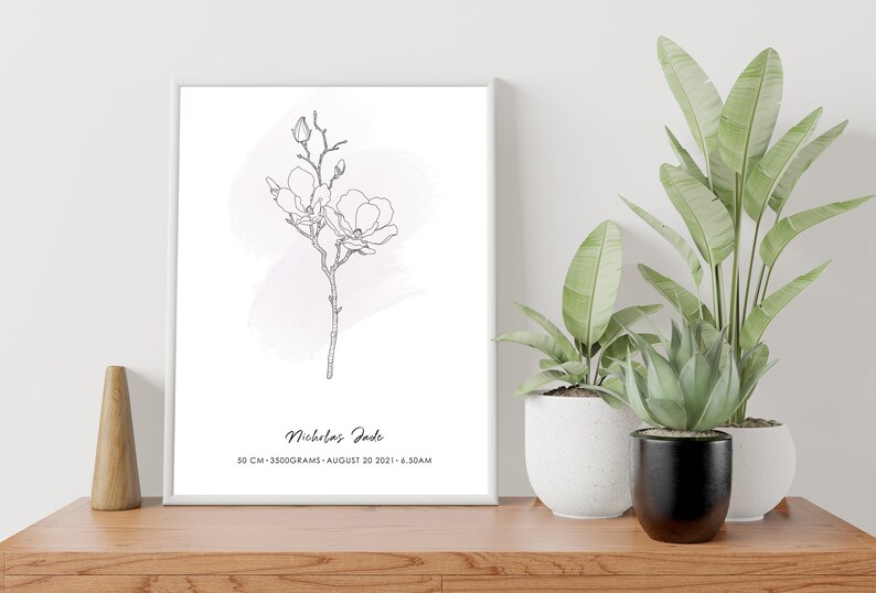 Puede incluir: Una impresi&oacute;n enmarcada con una ilustraci&oacute;n de flor de magnolia en blanco y negro y un suave lavado de acuarela rosa. La impresi&oacute;n incluye el nombre "Nicholas Jade", medidas y fecha. La impresi&oacute;n se muestra sobre una superficie de madera con plantas en macetas.
