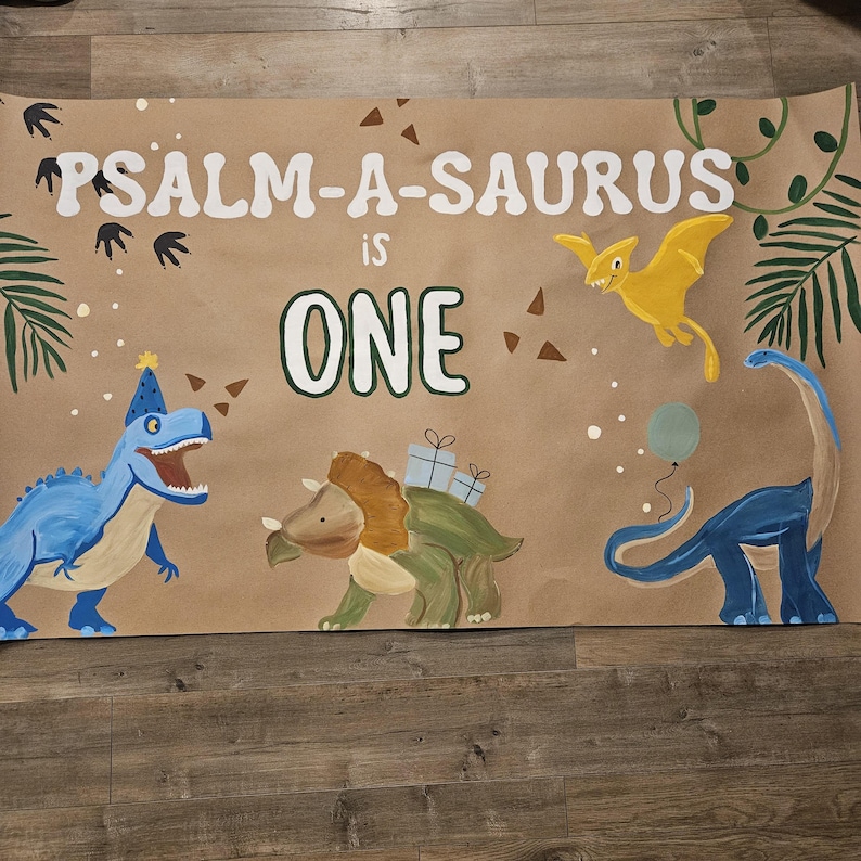 May include: A brown paper banner with the text "PSALM-A-SAURUS is ONE" in white paint. The banner is decorated with dinosaurs, including a blue dinosaur with a party hat, a green dinosaur with a brown head, a yellow flying dinosaur, and a blue dinosaur with a long neck. The banner is perfect for a first birthday party.
