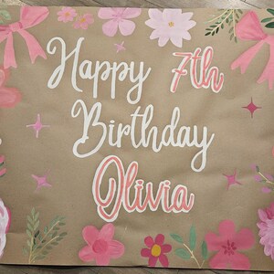 May include: A brown paper banner with the words "Happy 7th Birthday Olivia" written in white paint. The banner is decorated with pink flowers and bows.