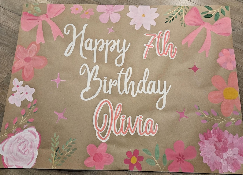 May include: A brown paper banner with the words "Happy 7th Birthday Olivia" written in white paint. The banner is decorated with pink flowers and bows.