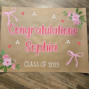 May include: A brown paper banner with the text "Congratulations Sophia Class of 2025" written in white paint. The banner is decorated with pink bows, flowers, and hearts.