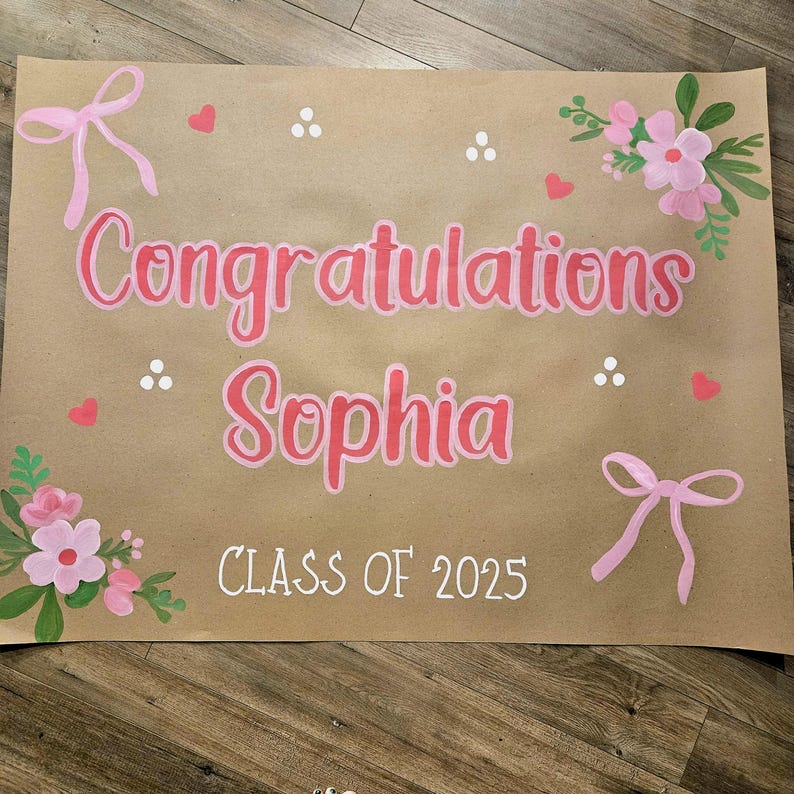 May include: A brown paper banner with the text "Congratulations Sophia Class of 2025" written in white paint. The banner is decorated with pink bows, flowers, and hearts.