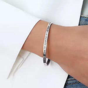 May include: A silver-colored bracelet with the German text "DENKE DARAN, DASS DU UNENDLICH VIEL KRAFT IN DIR HAST" engraved on the outside. The bracelet is worn on a wrist, with a white shirt and denim jeans in the background.