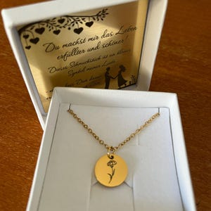 Könnte beinhalten: Goldfarbene Halskette mit rundem Anhänger mit schwarzem Blumendesign, präsentiert in einer weißen Geschenkbox. Die Box enthält eine goldfarbene Karte mit deutschem Text und einer Paarsilhouette.