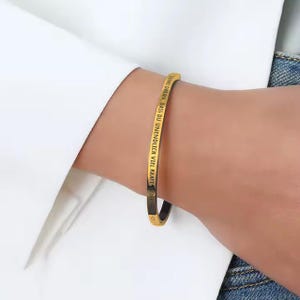 Può includere: Un bracciale rigido color oro con testo nero. Il bracciale è indossato su un polso e il testo sembra essere una citazione motivazionale in tedesco. Il bracciale ha un design semplice ed elegante.