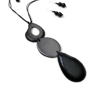 Puede incluir: Un collar negro con un colgante hecho de tres círculos de madera negra. El collar está hecho de cordón negro y tiene un diseño simple. El colgante está hecho de tres círculos, uno grande, uno mediano y uno pequeño. Los círculos están todos conectados por un cordón negro. El collar es una pieza de joyería simple y elegante.