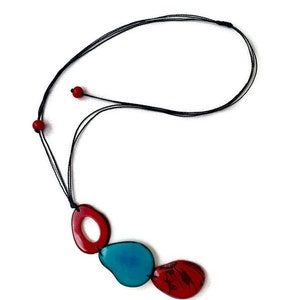 May include: A necklace with a black cord and three pendant charms. The charms are shaped like an oval, a teardrop, and a rounded rectangle. The oval and rectangle charms are red, and the teardrop charm is blue.