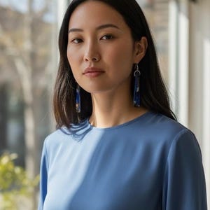 May include: A woman wearing blue earrings and a blue long-sleeve top. The earrings are long, rectangular, and feature a blue and gold pattern. The top is a solid, light blue color.