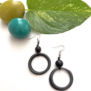 May include: A pair of black hoop earrings with a small black bead above each hoop. The earrings are displayed with a green leaf, a teal sphere, and a yellow-green fruit against a white background. The earrings have silver-colored ear wires.
