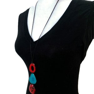 May include: A long black cord necklace with three ceramic pendant charms. The charms are red, blue, and green. The necklace is worn on a black shirt.