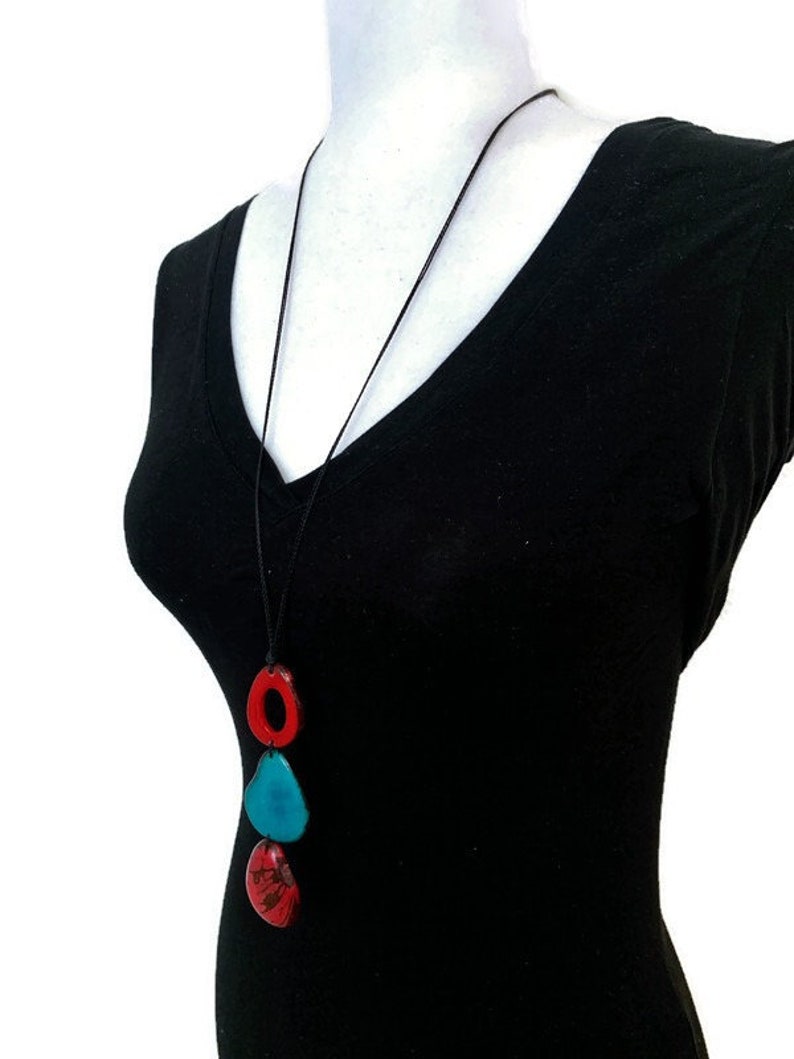 May include: A long black cord necklace with three ceramic pendant charms. The charms are red, blue, and green. The necklace is worn on a black shirt.