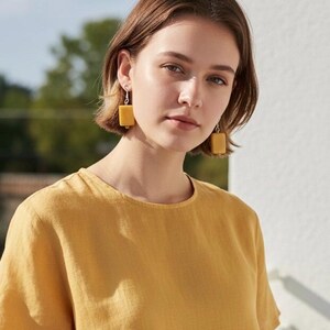 May include: A woman wearing yellow square earrings and a yellow top. The earrings are a solid, muted yellow color and hang from silver hooks. The top is a simple, short-sleeved design in a matching yellow hue.