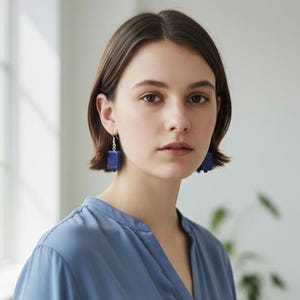 May include: A woman wearing blue square drop earrings. The earrings are a deep blue color and hang from silver hooks. She is wearing a blue blouse. The background is a soft, neutral color.