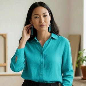 May include: A woman wearing a turquoise button-down shirt and black pants. She is also wearing turquoise earrings. The shirt has long sleeves with a buttoned cuff. The background is a neutral color.
