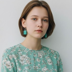 Puede incluir: Una persona con pendientes rectangulares turquesas. Los pendientes están suspendidos de ganchos plateados. Lleva un vestido verde claro con estampado floral. La persona tiene el pelo corto castaño y mira a la cámara.