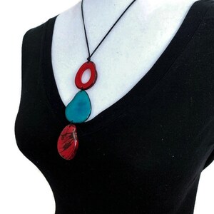 May include: A necklace with three colorful, abstract, resin pendants. The top pendant is a red ring with a smaller red ring inside. The middle pendant is a teal teardrop shape. The bottom pendant is a red teardrop shape with a marbled pattern.