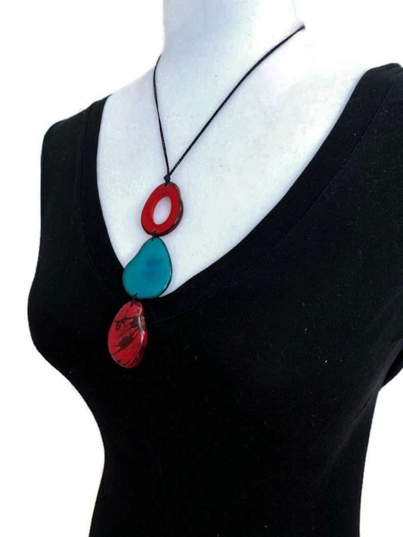 May include: A necklace with three colorful, abstract, resin pendants. The top pendant is a red ring with a smaller red ring inside. The middle pendant is a teal teardrop shape. The bottom pendant is a red teardrop shape with a marbled pattern.