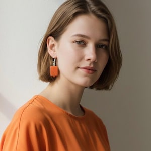 Op de afbeelding: Een persoon met oranje vierkante oorbellen. De oorbellen hangen aan zilveren haken. Ze draagt ook een oranje top. De persoon heeft kort bruin haar en kijkt in de camera.
