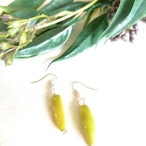 May include: A pair of handmade earrings featuring elongated, light green beads with silver-colored hooks. The earrings are displayed against a white background, with green leaves and buds in the upper portion of the image. The earrings are a simple, elegant design.